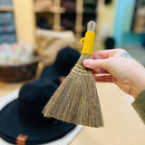 A Hand holding up a small mini hand-broom with yellow wrapped string for the handle. This is a Free Gift with every Packable Ash Hat Purchase for the month of October and Spooky season