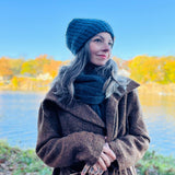 Woman wearing a brown coat and blue knit hat by a lake with autumn foliage.
