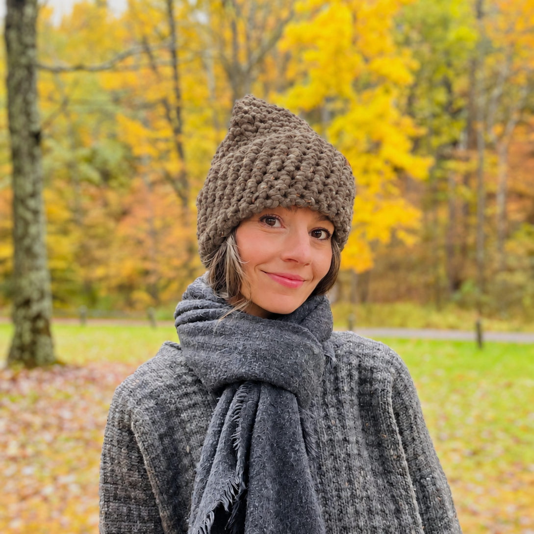 A woman wearing a North Ferry Hat handmade in the Finger Lakes, fits Men Women and Kid Sizes Unisex Hatschunky brown knit beanie and a dark gray waffle-knit scarf and sweater, standing outdoors in a park with autumn trees in the background.