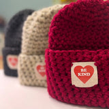 Three knitted hats in different colors with 'Be Kind' patches on a light background