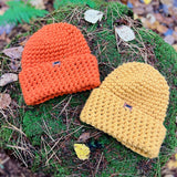 Two knitted beanies, one orange and one yellow, on a mossy log with a natural background.