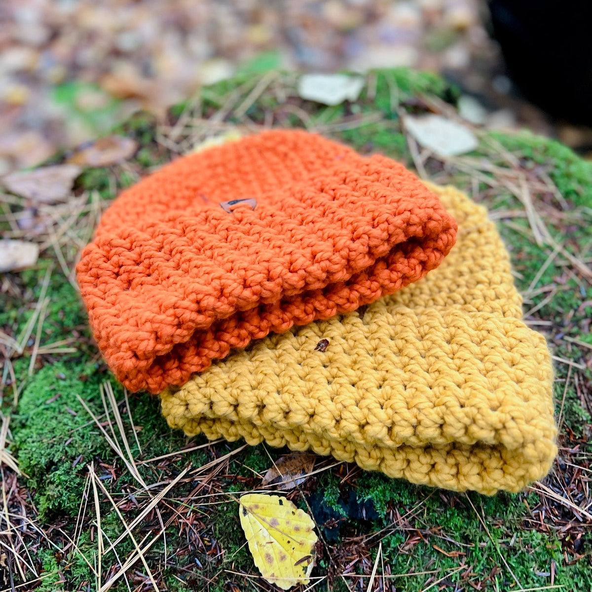 Two crocheted beanies, one orange and one yellow, on a mossy surface with fallen leaves.