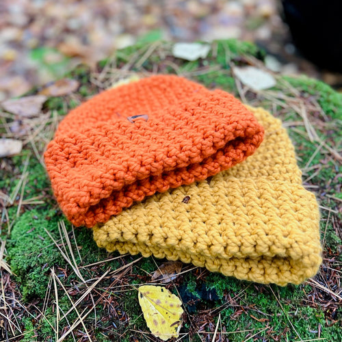 Two crocheted beanies, one orange and one yellow, on a mossy surface with fallen leaves.