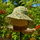 A wide-brim sun hat with a green and white leafy camouflage pattern, displayed on a wooden stand outdoors in a garden with tomato plants and green foliage.