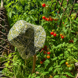 A wide-brimmed bucket hat with a green, beige, and black geometric pattern is displayed outdoors in a garden among tomato plants and trellis.