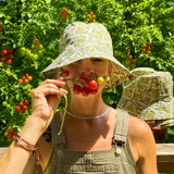 A woman outdoors in a garden wearing a patterned bucket hat and olive green overalls, holding a cluster of red and yellow berries near her face. 