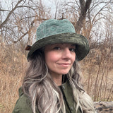 Person wearing a green hat and coat in a natural setting wearing a Handmade North Ferry Bucket Hat that is reversible, waterproof, relaxed fit style