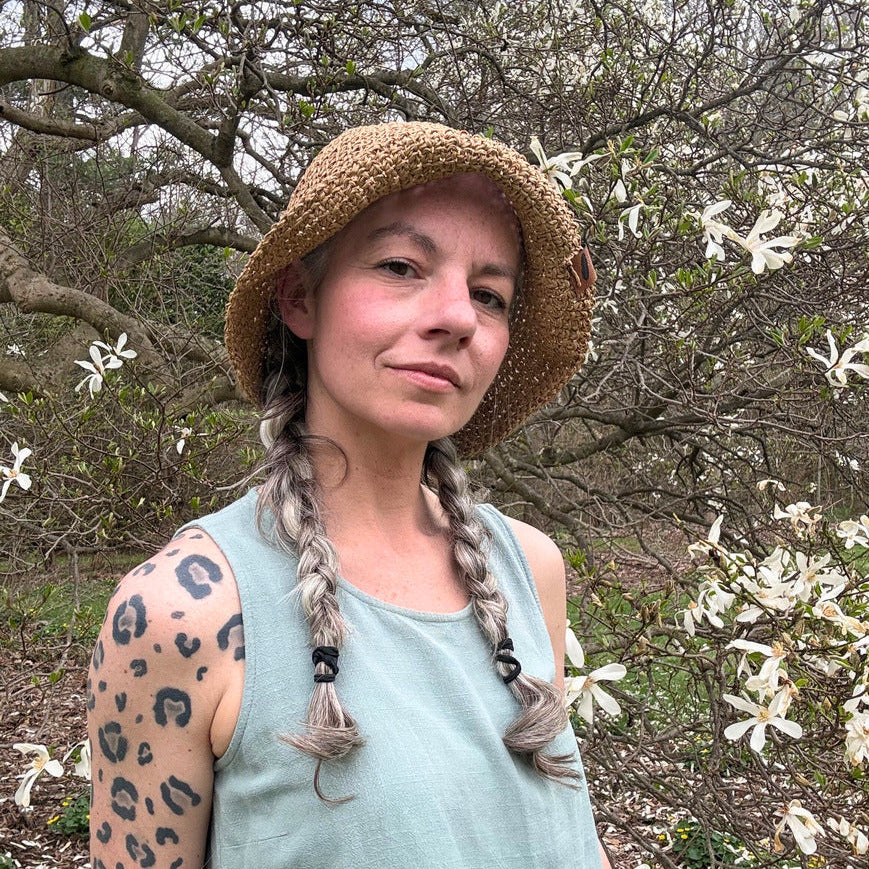 Woman wearing a straw hat and light blue top standing in a natural setting with trees and flowers. Floppy Flexible Brims for Packable Straw Hats
