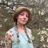 Woman wearing a straw hat and light blue top standing in a natural setting with trees and flowers. Floppy Flexible Brims for Packable Straw Hats